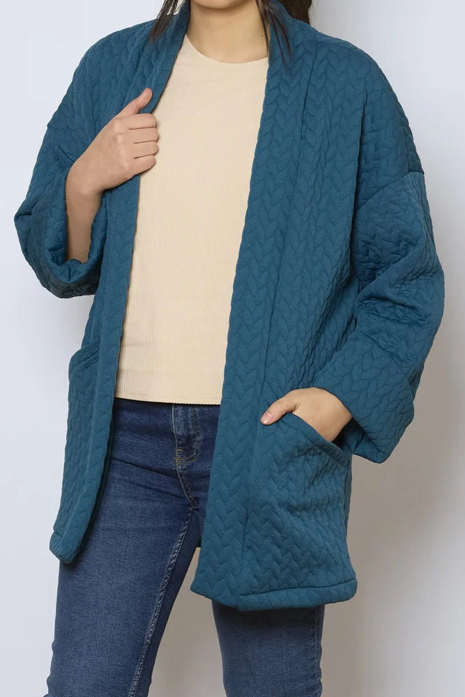 A woman wearing the Ina Coatigan by Tangente in Peacock, an open front cardigan in a cable-knit textured fabric, with dropped sleeves and patch pockets. She is wearing it with a white shirt and jeans and standing in front of a white background.