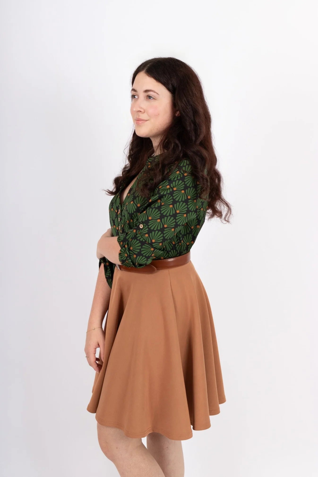 Side view of a women wearing the Leonie Blouse by Infime in Black with Green print, featuring a notched V-neckline, three button placket, and 3/4 sleeves. She is wearing it with a tan skirt and standing in front of a white background. 