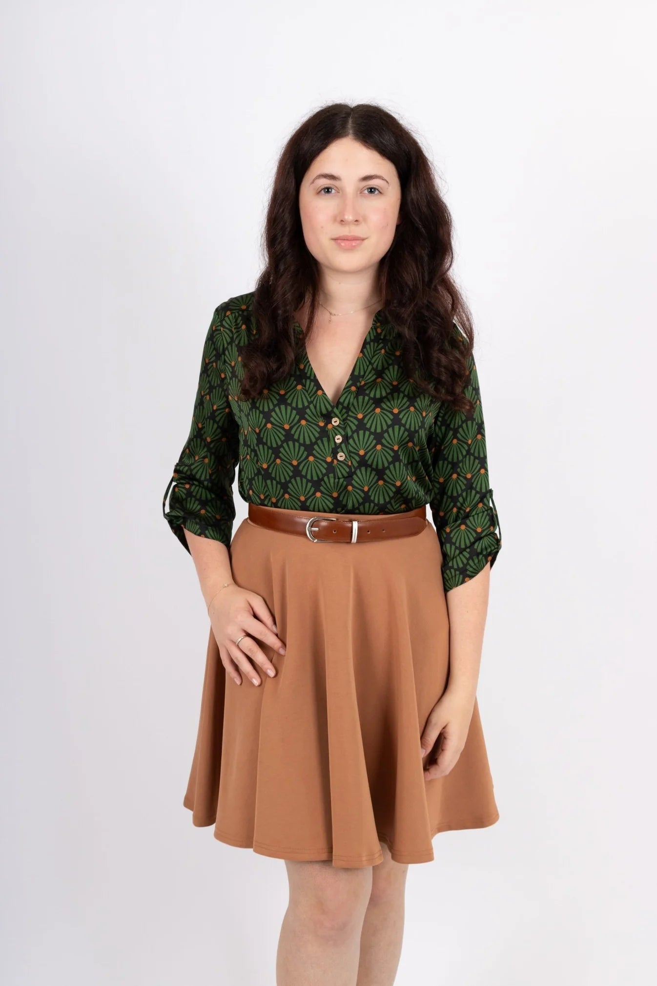 A woman wearing the Leonie Blouse by Infime in Black with Green print, featuring a notched V-neckline, three button placket, and 3/4 sleeves. She is wearing it with a tan skirt and standing in front of a white background. 