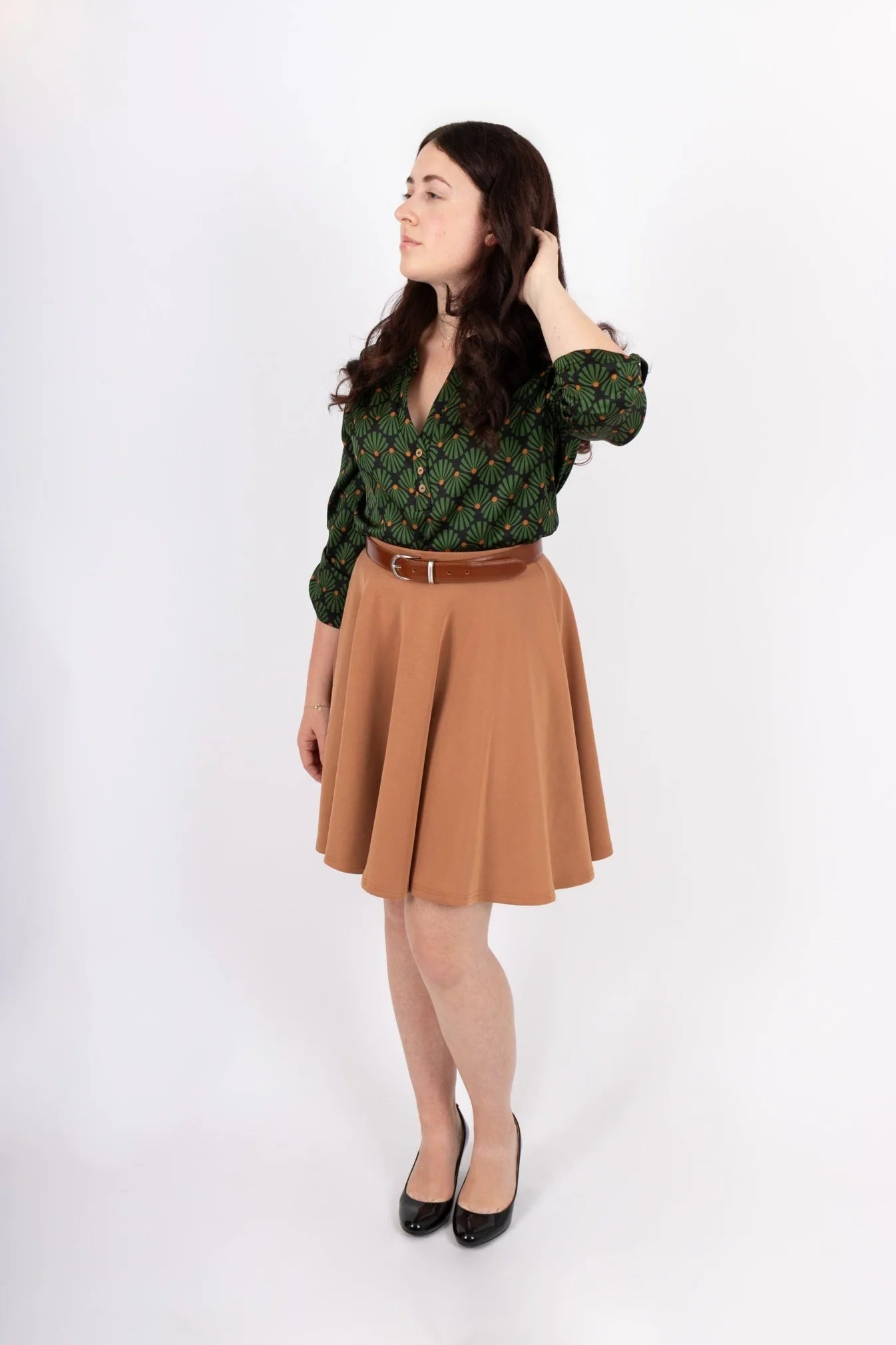 A women wearing the Leonie Blouse by Infime in Black with Green print, featuring a notched V-neckline, three button placket, and 3/4 sleeves. She is wearing it with a tan skirt and standing in front of a white background. 