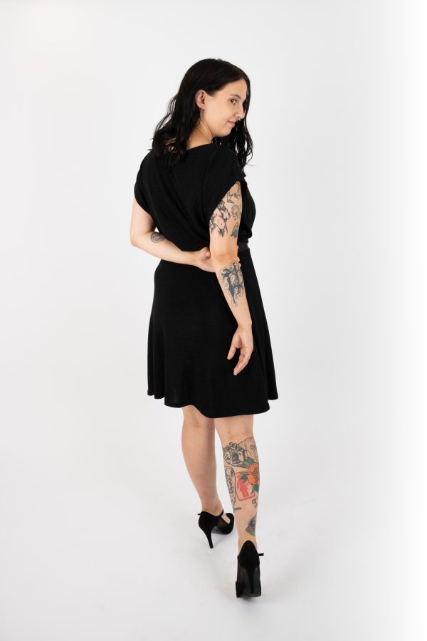 Back view of A woman wearing the Miyuki Dress by Infime in Black, with glittery fabric, a boat neck, short extended sleeves, a contrast waistband, and a flared knee-length skirt, standing in front of a white wall