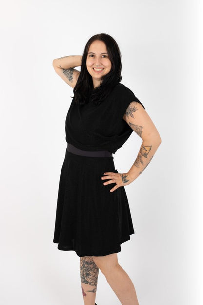 A woman wearing the Miyuki Dress by Infime in Black, with  glittery fabric, a boat neck, short extended sleeves, a contrast waistband, and a flared knee-length skirt, standing in front of a white background 