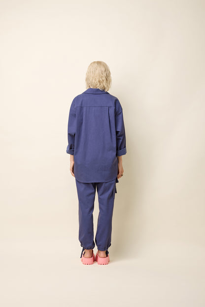 Back view of a woman wearing the Iyrna Shirt by Cokluch in Twiight blue, an oversized long sleeved button front shirt with chest flap pocket with a ring closure detail. She is wearing it with matching pants and standing in front of a beige background.  