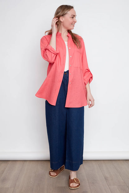 A woman wearing the jasmine Long Sleeve Shirt by Julei in Coral, a classic dress shirt style with a mandarin collar, pleated pocket, and cuffed sleeves. She is wearing it with jeans and standing in front of white background. 