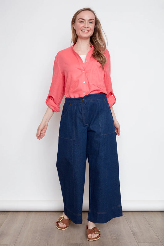 A woman wearing the jasmine Long Sleeve Shirt by Julei in Coral, a classic dress shirt style with a mandarin collar, pleated pocket, and cuffed sleeves. She is wearing it with jeans and standing in front of white background. 