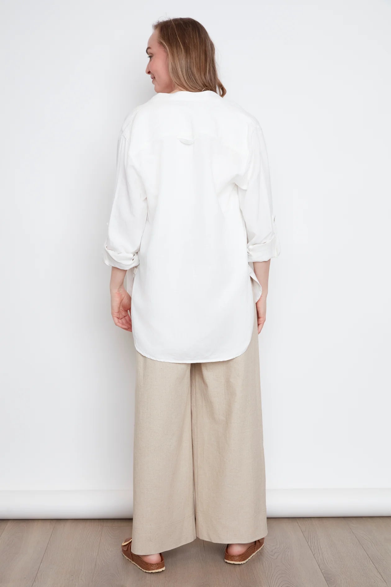 Back view of a woman wearing the jasmine Long Sleeve Shirt by Julei in Natural White, a classic dress shirt style with a mandarin collar, pleated pocket, and cuffed sleeves. She is wearing it with jeans and standing in front of white background. 