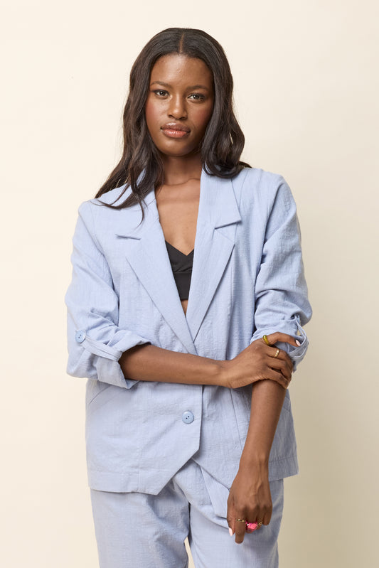 A woman wearing the Jennifer Jacket by Cokluch in Dove, with a wide lapel collar, two front buttons, angled front helm, and adjustable sleeves. She is wearing it with matching pants and standing in front of a beige background. 