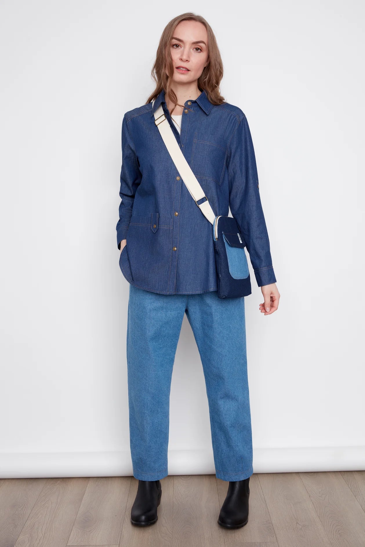 A woman wearing the Jasmine Denim Shirt by Julei, a boyfriend style button front shirt with a chest pocket and a large front patch pocket. She is wearing it with jeans and standing in front of a white background. 