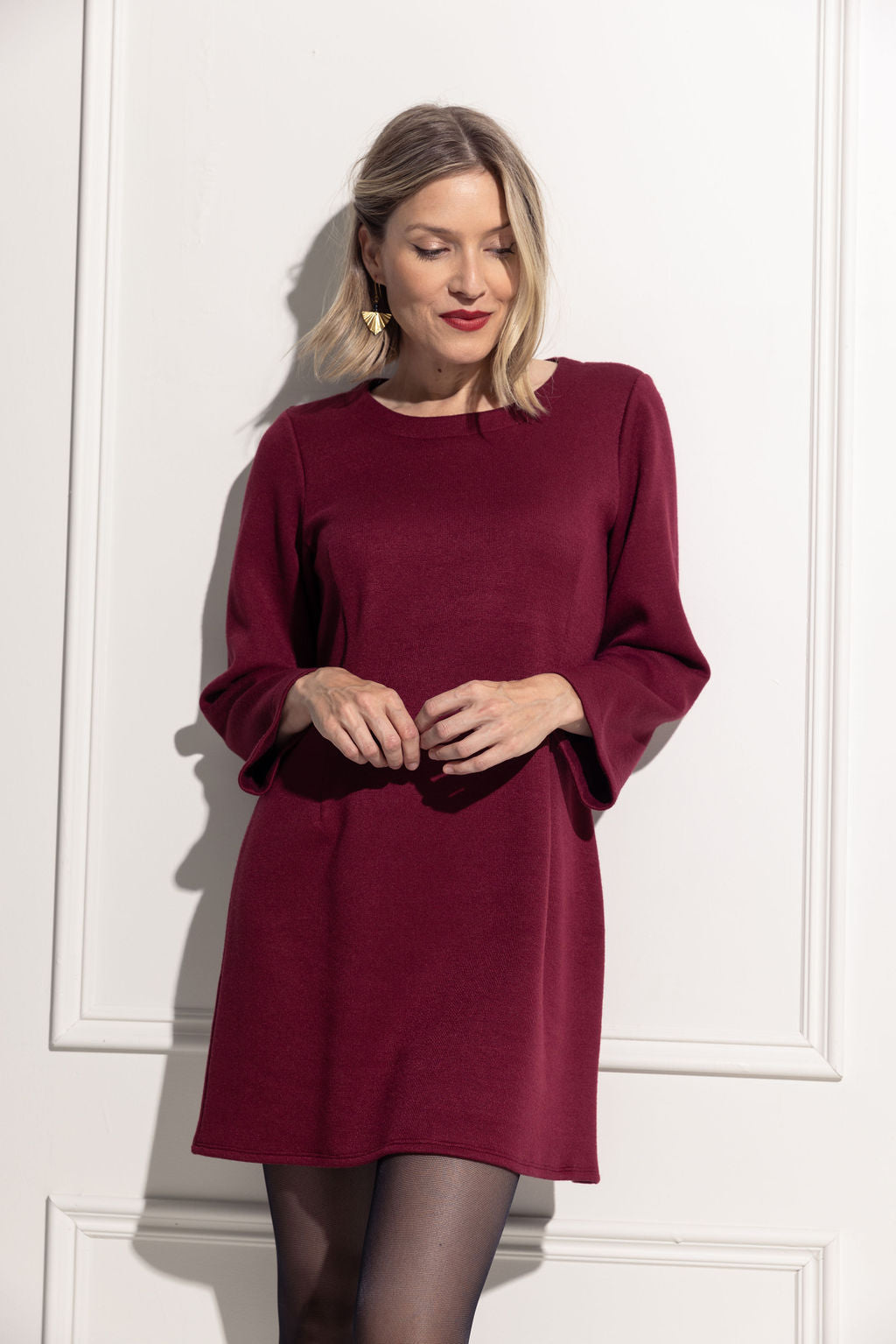 A woman wearing the Blondie Dress by Cherry Bobin in Red, made from eco-friendly knit fabric, with a round neck, long sleeves, darts down the front, and a mid-thigh hem. She is standing in front of a white wall. 