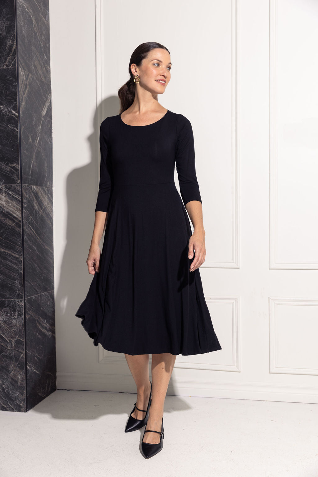 A woman wearing the Diamond Dress by Cherry Bobin in Black, featuring a boat neck, 3/4 sleeves, a fit and flare shape, and a tea-length skirt in bamboo rayon. She is standing. in front of a white wall. 