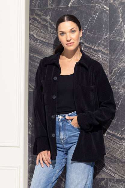 A woman wearing the Joplin Jacket by Cherry Bobin in Black, a shirt jacket in corduroy fabric with a collar and button front and two breast pockets. She is wearing it over jeans and a black shirt and is standing in front of a grey wall. 