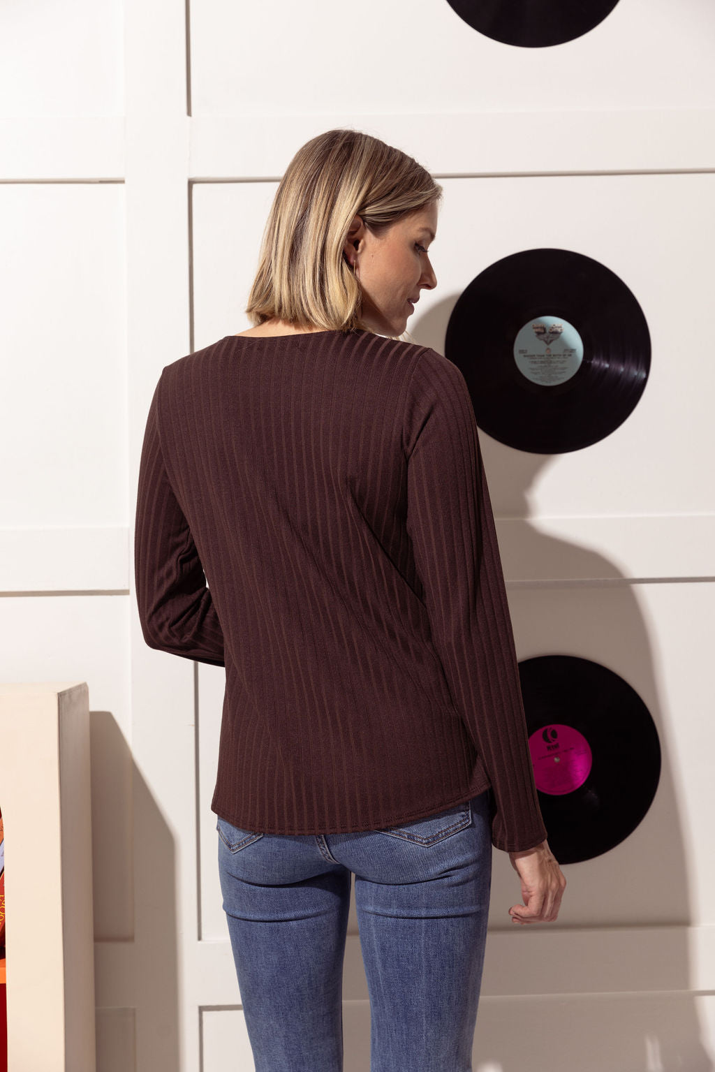 Back view of a woman wearing the Silver Sweater by Cherry Bobin in Brown, with a round collar, long sleeves, slim fit, and wide rib knit, with jeans, standing in front of a wall with vinyl records hanging on it. 