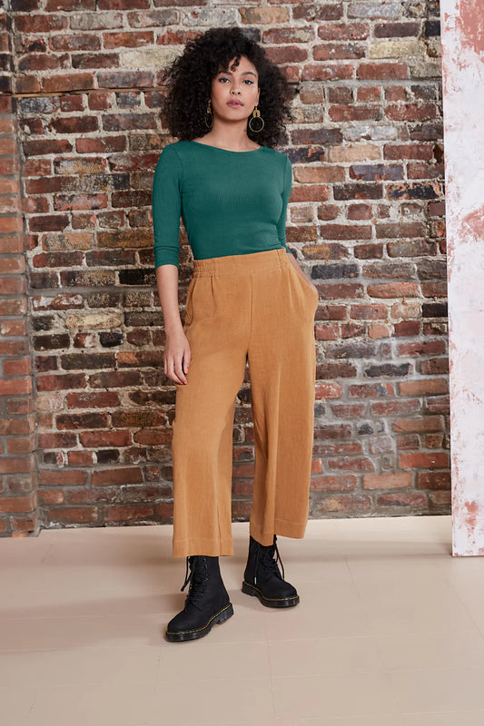 A woman wearing the Kurt Pants by Melow in Honey, featuring wide cropped legs, a flat-front elastic waist band, and slant pockets.  She is wearing them with a green top and standing in front of a brick wall. 