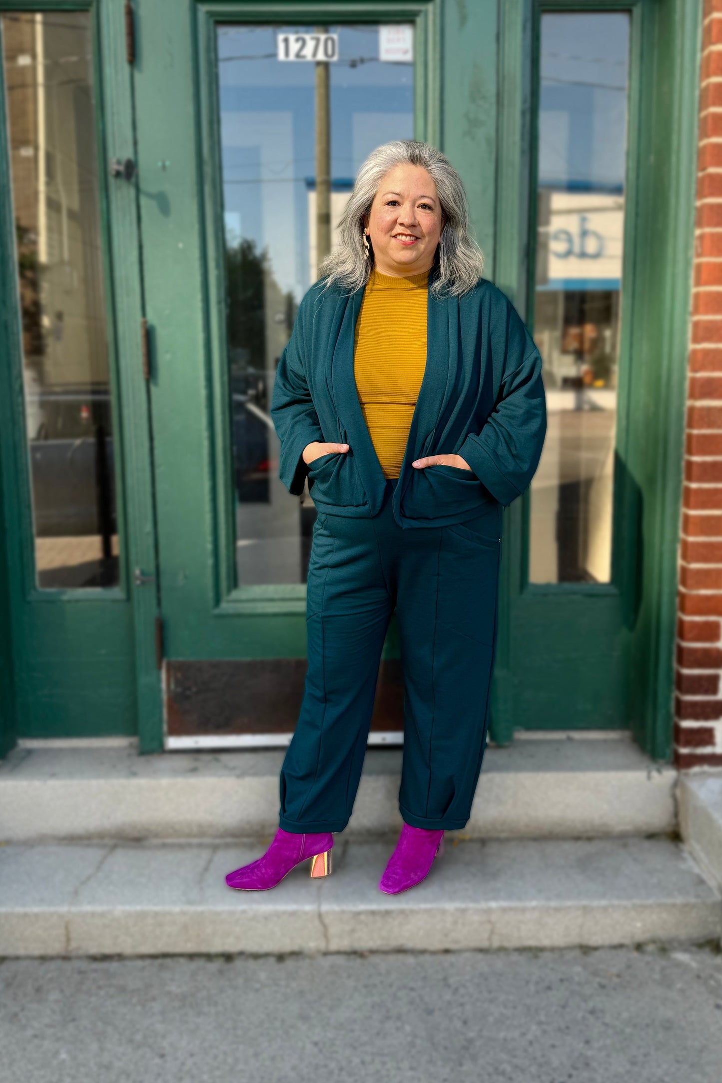 Christina wearing the Herbst Pant and Thuya Jacket by Kazak in Teal, standing in front of a green door 