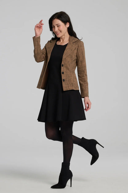 A woman wearing the Toscana Dress by Kollontai in Black, a sleeveless piece with a round neck, princess seams, and fit and flare shape with a full skirt that hits above the knee. She is wearing it with a brown blazer and standing against a grey background.