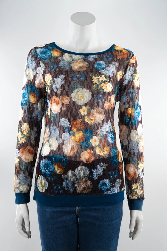 The Aubade Lace Top by Karkass, featuring a floral print on a brown background, round neck, and long sleeves with contrasting teal at the collar, cuffs, and hem, is shown on a mannequin with jeans in front of a white background. 