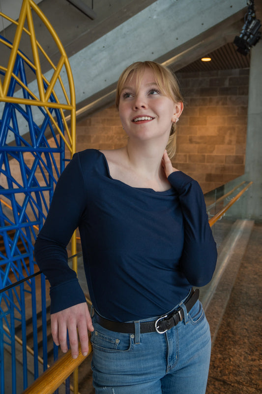 A woman wearing the Drapé Top by Karkass in Navy, a long sleeved rayon jersey top with a draped boatneck and a slim fit. she is standing indoors in front of a colourful structure. 