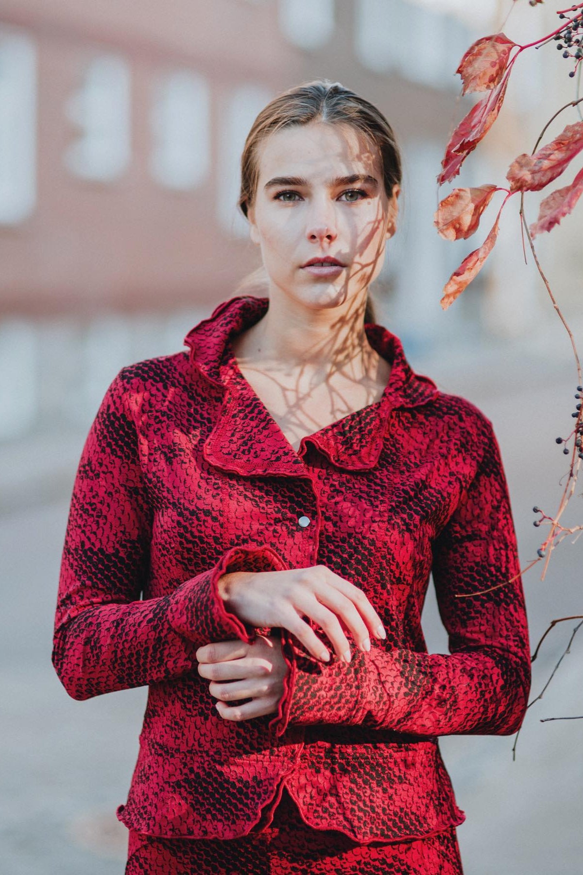 A woman wearing the Essential Jacket by Karkass in Red Snakeskin, a fitted jacket with a ruffled collar, button front, and frills at the hem and cuffs. She is wearing it with matching pants and standing on a street. 