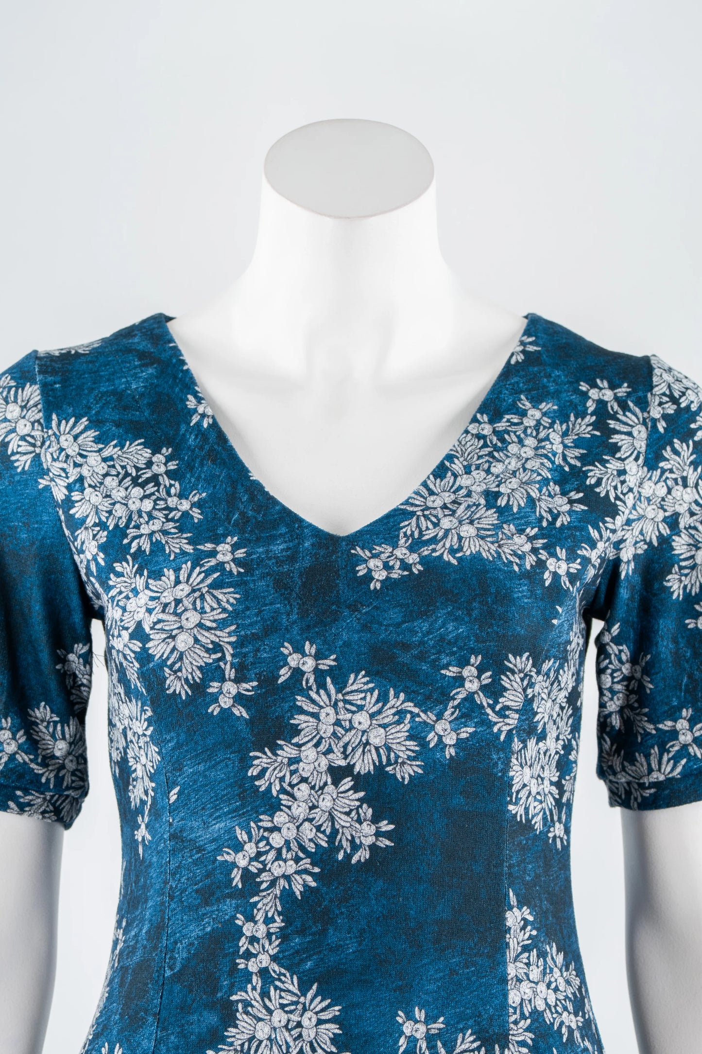 The Ice Flower Dress by Karkass in Blue Floral, a knit dress with a V-neck, elbow length sleeves, fit and flare shape, and above the knee hem, is shown on a mannequin in front of a white background. 