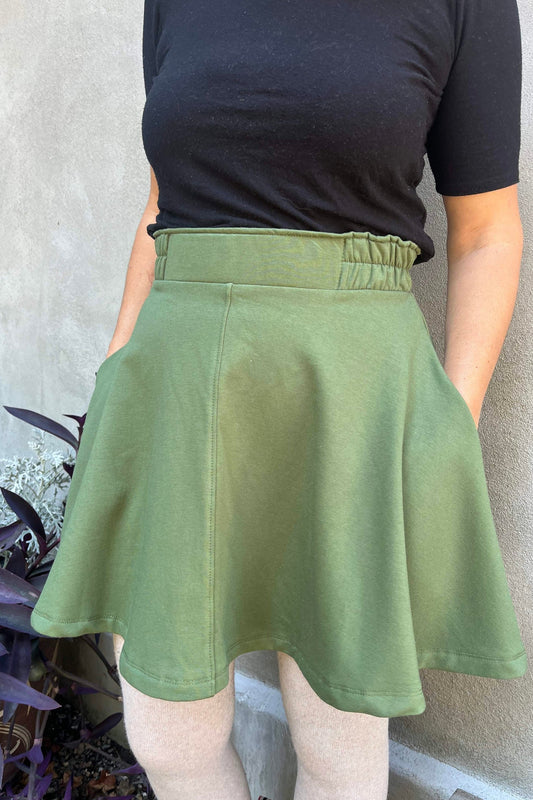 Close up detail of a woman wearing the Juniper Skirt by Kazak in Olive, a full circle skirt with an elastic waist, above the knee length, and side pockets. She is wearing it with a black top and standing in outside in front of a wall. 