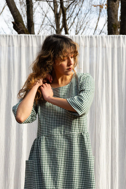 A woman wearing the Persea Dress by Kazak in Forest Gingham print, a below-the-knee length dress with 3/4 sleeves, a removable belt, and large patch pockets. She is standing in a forest in front of a white sheet. 