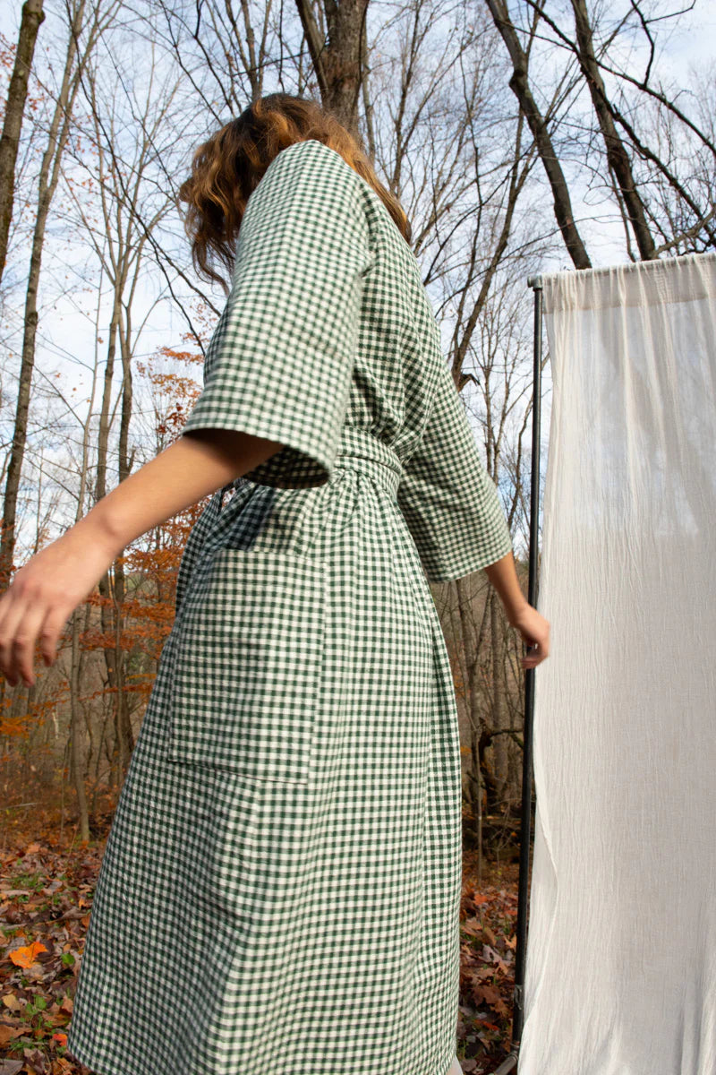 A woman wearing the Persea Dress by Kazak in Forest Vichy print, a below-the-knee length dress with 3/4 sleeves, a removable belt, and large patch pockets. She is standing in a forest in front of a white sheet. 