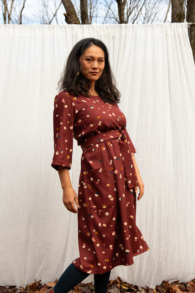 A woman wearing the Persea Dress by Kazak in Bordeaux Moon print, a below-the-knee length dress with 3/4 sleeves, a removable belt, and large patch pockets. She is standing in an autumn forest in front of a white curtain. 