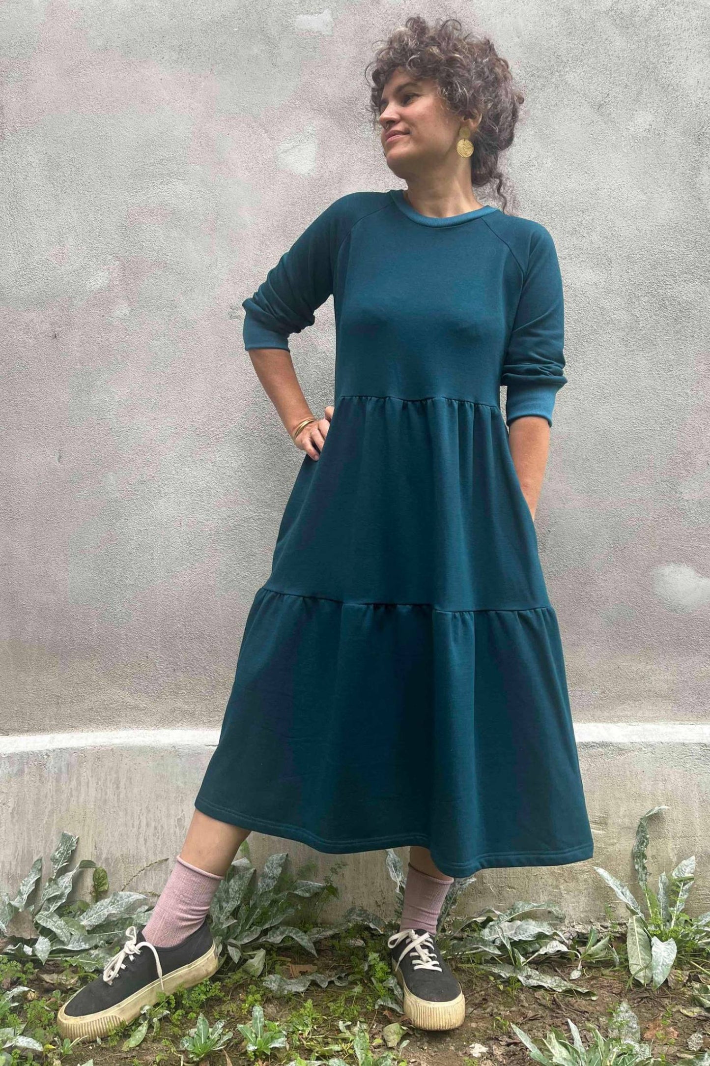 A woman wearing the Simone Dress by Kazak in Teal, made from organic cotton and featuring a round neck, 3/4 raglan sleeves, and a tiered midi-length skirt. She is standing outside on front of a grey wall. 