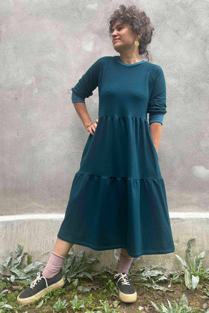 A woman wearing the Simone Dress by Kazak in Teal, made from organic cotton and featuring a round neck, 3/4 raglan sleeves, and a tiered midi-length skirt. She is standing outside on front of a grey wall. 