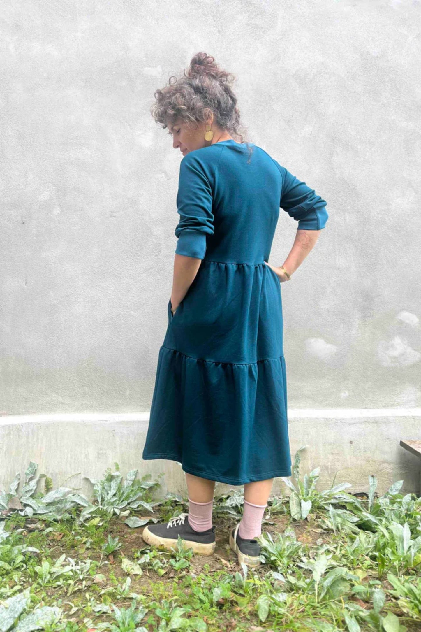 Back view of a woman wearing the Simone Dress by Kazak in Teal, made from organic cotton and featuring a round neck, 3/4 raglan sleeves, and a tiered midi-length skirt. She is standing outside on front of a grey wall. 