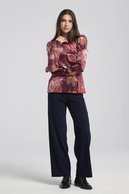 A woman wearing the Hermione Pants by Kollontai in Navy, stretchy corduroy pants with a wide pull-one waistband, wide legs, and angled patch pockets. She is wearing them with patterned burgundy top and standing in front of a grey background. 