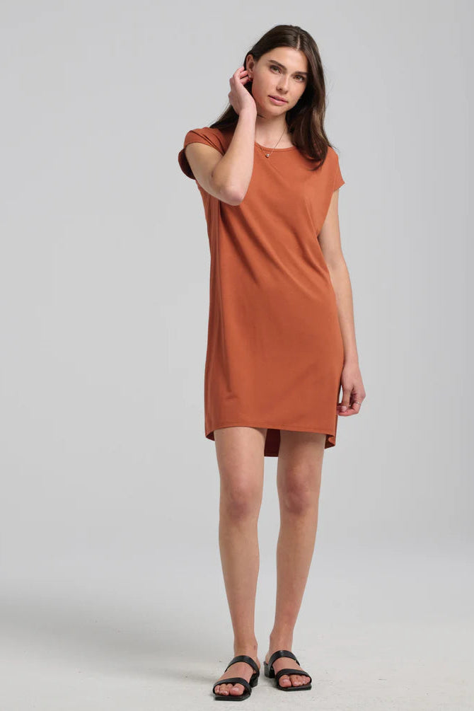 A woman wearing the Arwen Dress by Kollontai in Black, a t-shirt dress with a round neck, short extended sleeves, a loose ft, and an above the knee hem. She is standing in front of a grey background. 