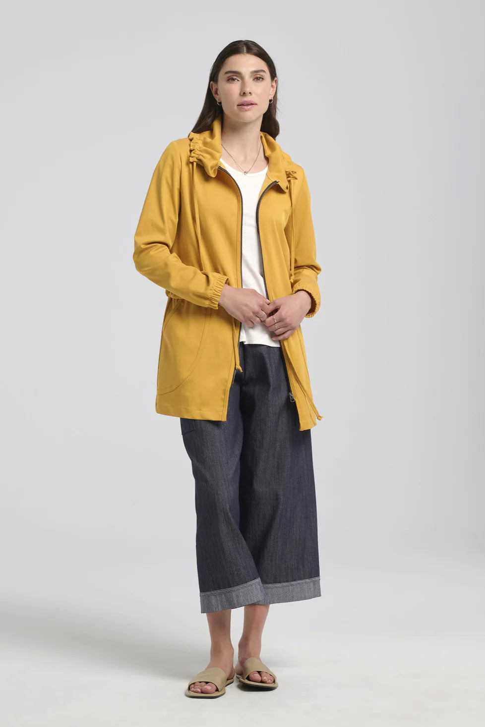 A woman wearing the Bronwen Jacket by Kollontai in Mustard, a mid-thigh length jacket with drawstrings at the waist and hood and large patch  pockets. She is wearing it with the Morwenna Pants and standing in front of a white background. 