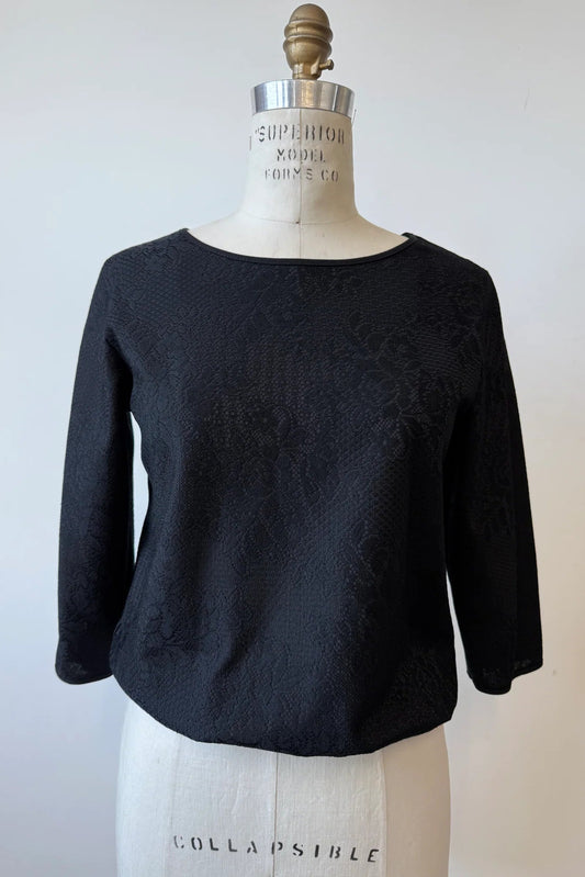 The Celestine Blouse By Kollontai in Black, featuring black satin overlaid with black lace, is shown on a mannequin in front of a white background. It has a cropped silhouette with an elastic waist, and 3/4 sleeves. 