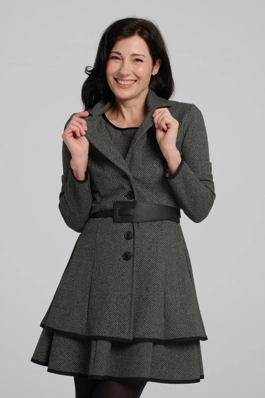A woman wearing the Henderson Jacket by Kollontai in Charcoal tweed, with a fitted A-line shape that flares from the waist, a classic collar, and buttons down the front. She is wearing it with a matching dress and standing in front of a grey background. 