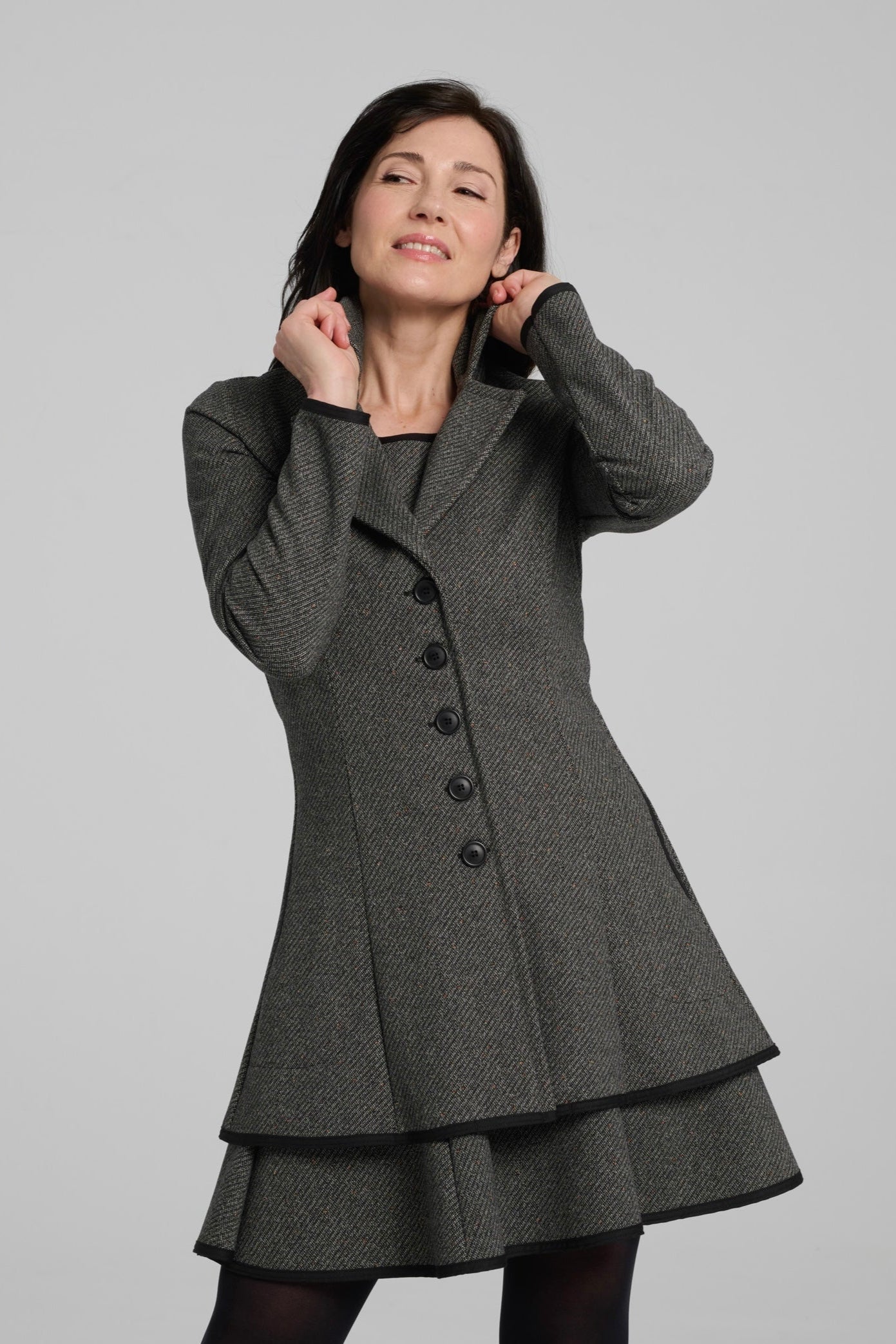 A woman wearing the Henderson Jacket by Kollontai in Charcoal tweed, with a fitted A-line shape that flares from the waist, a classic collar, and buttons down the front. She is wearing it with a matching dress and standing in front of a grey background. 