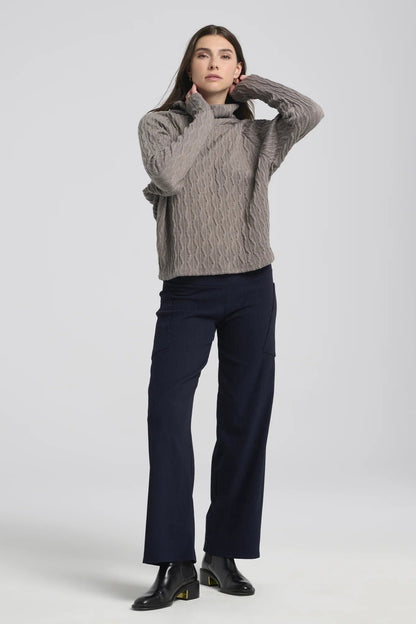 A woman wearing the Hermione Pants by Kollontai in Navy, stretchy corduroy pants with a wide pull-one waistband, wide legs, and angled patch pockets. She is wearing them with a grey sweater and standing in front of a grey background. 
