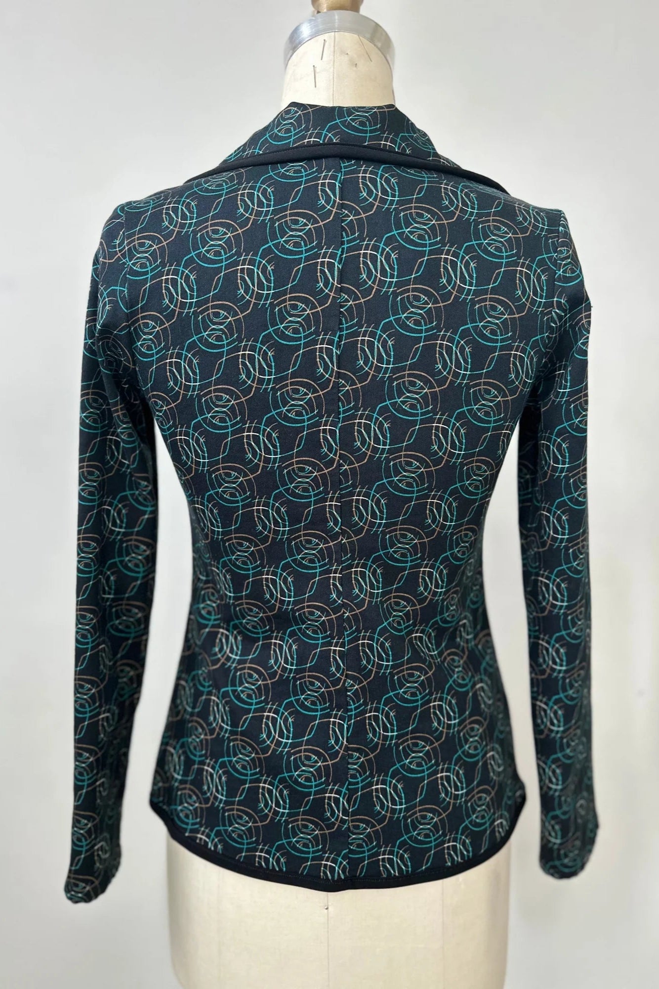 A back view of the Nara Jacket by Kollontai in Turquoise, a blazer with a turquoise on black geometric print, with a rounded lapel collar, slim fit, and side pockets, is shown on a mannequin in front of a grey background. 