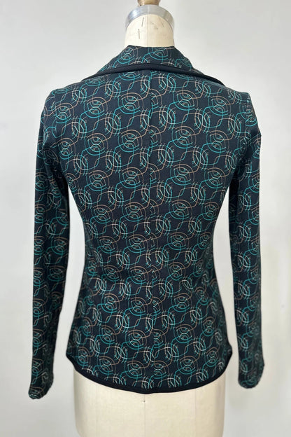 A back view of the Nara Jacket by Kollontai in Turquoise, a blazer with a turquoise on black geometric print, with a rounded lapel collar, slim fit, and side pockets, is shown on a mannequin in front of a grey background. 