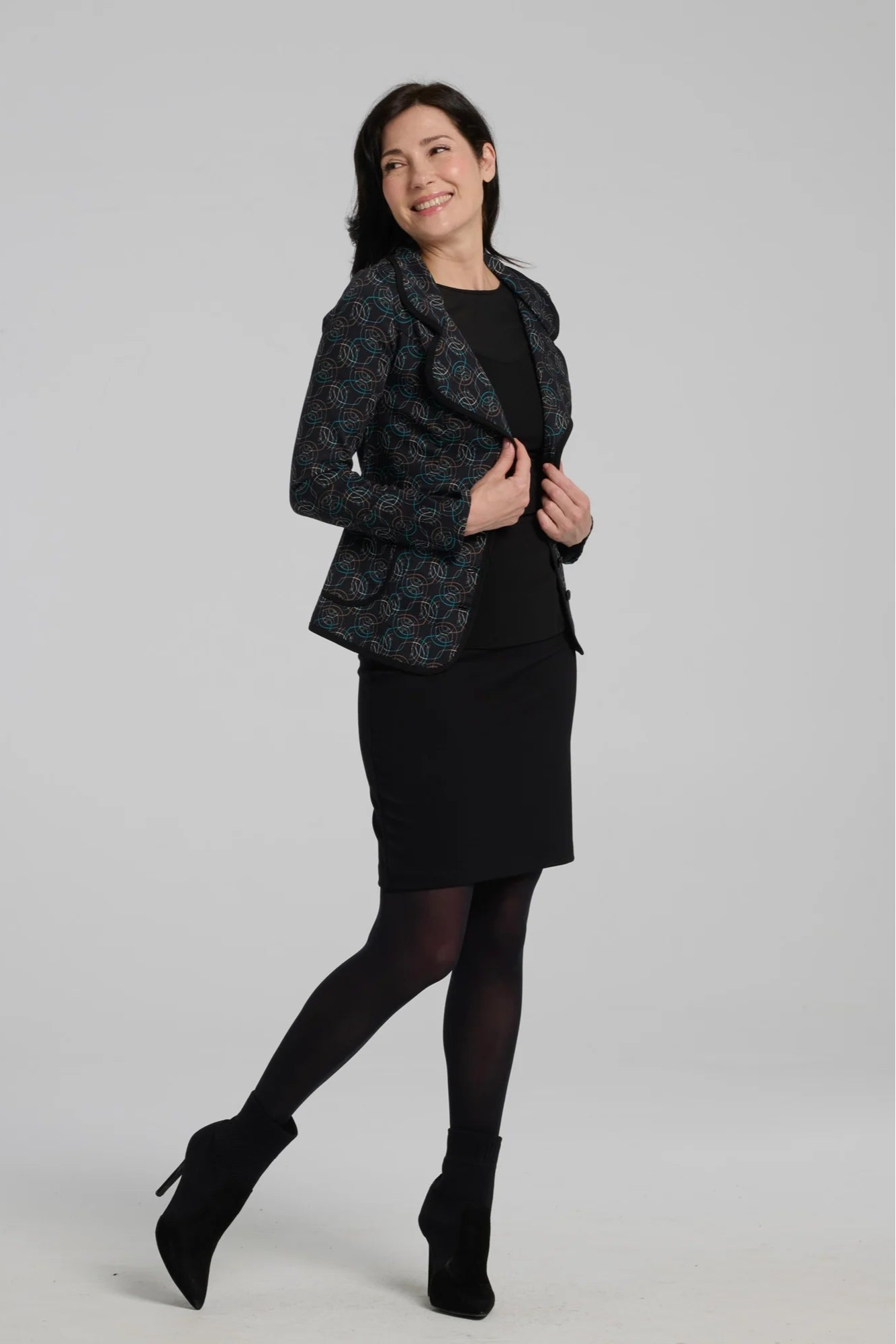 A woman wearing the Nara Jacket by Kollontai in Turquoise, a blazer with a turquoise on black geometric print, wit a rounded lapel collar, slim fit, and side pockets. She is wearing it with a black top and a black skirt and standing in front of a grey background. 