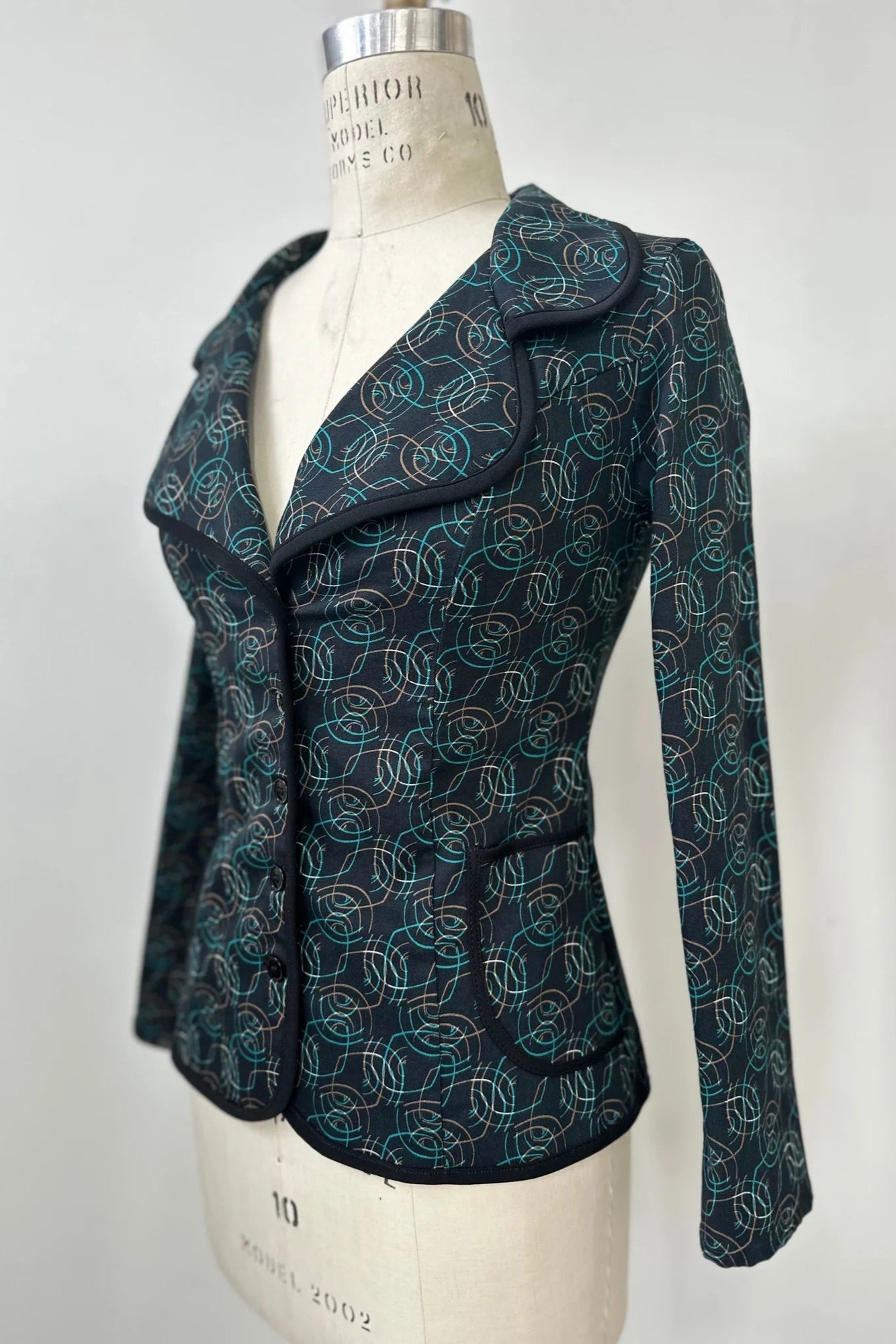 The Nara Jacket by Kollontai in Turquoise, a blazer with a turquoise on black geometric print, with a rounded lapel collar, slim fit, and side pockets, is shown on a mannequin in front of a grey background. 