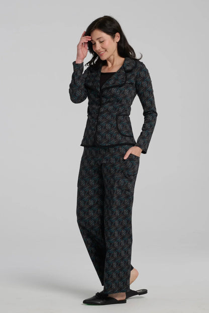 A woman wearing the Nara Jacket by Kollontai in Turquoise, a blazer with a turquoise on black geometric print, wit a rounded lapel collar, slim fit, and side pockets. She is wearing it with a black top and the matching Elma Pant and standing in front of a grey background. 