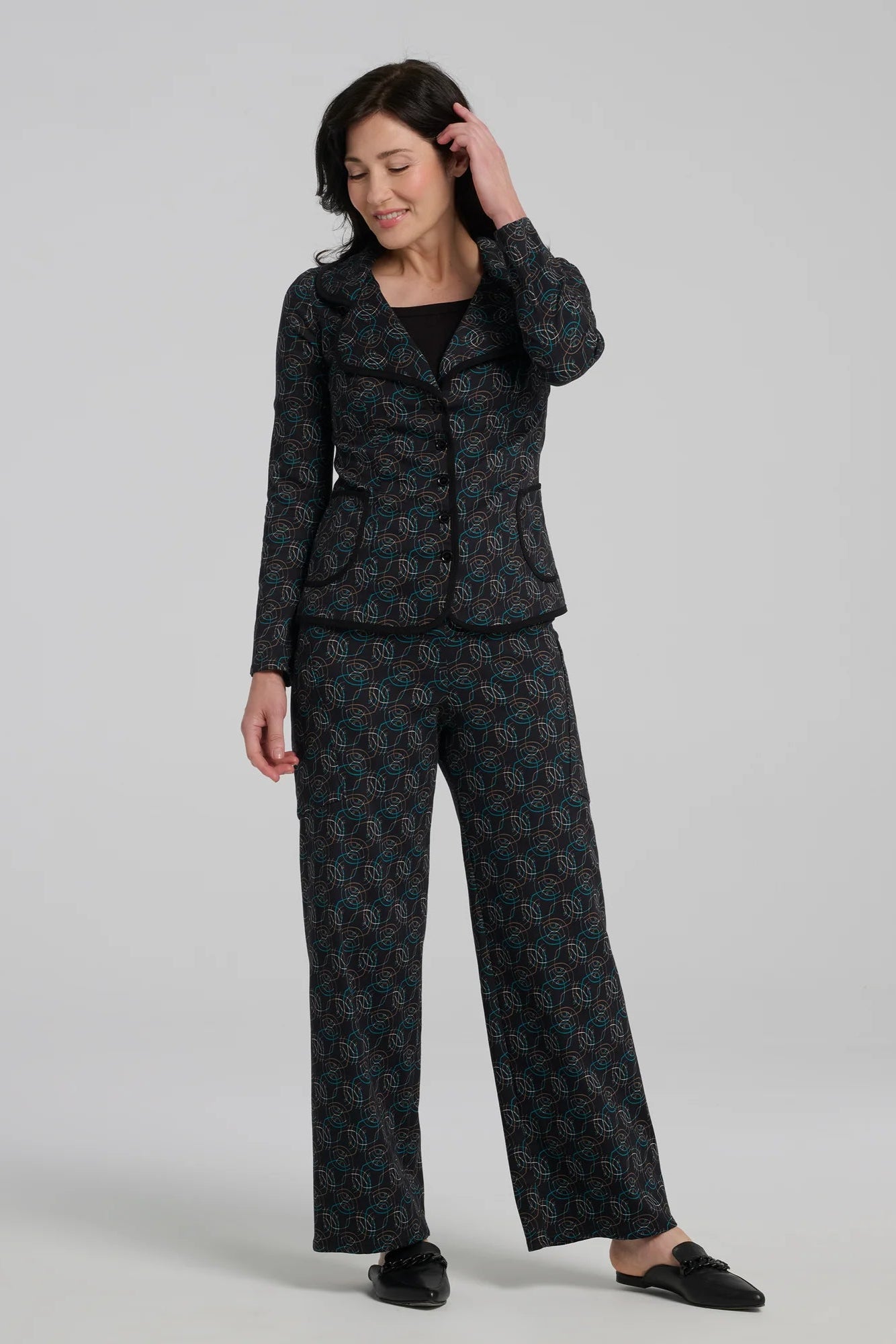 A woman wearing the Nara Jacket by Kollontai in Turquoise, a blazer with a turquoise on black geometric print, wit a rounded lapel collar, slim fit, and side pockets. She is wearing it with a black top and the matching Elma Pant and standing in front of a grey background. 