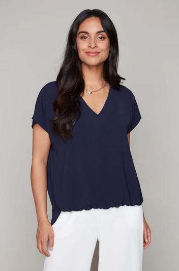 A woman wearing the Oakley Woven Top by Compli K in Navy, a short sleeved, loose fitting top with a V-neck. She's wearing it with white pants and sgtanding in front of a beige background. 