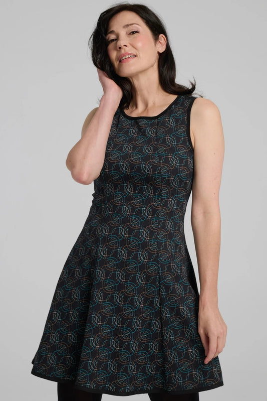 A woman wearing the Silba Dress by Kollontai in a Turquoise on black print, a sleeveless dress with a round neck, fit and flare shape, and above the knee hem She is standing against a grey background. 