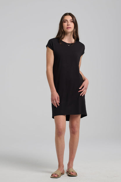 A woman wearing the Arwen Dress by Kollontai in Black, a t-shirt dress with a round neck, short extended sleeves, a loose ft, and an above the knee hem. She is standing in front of a grey background. 