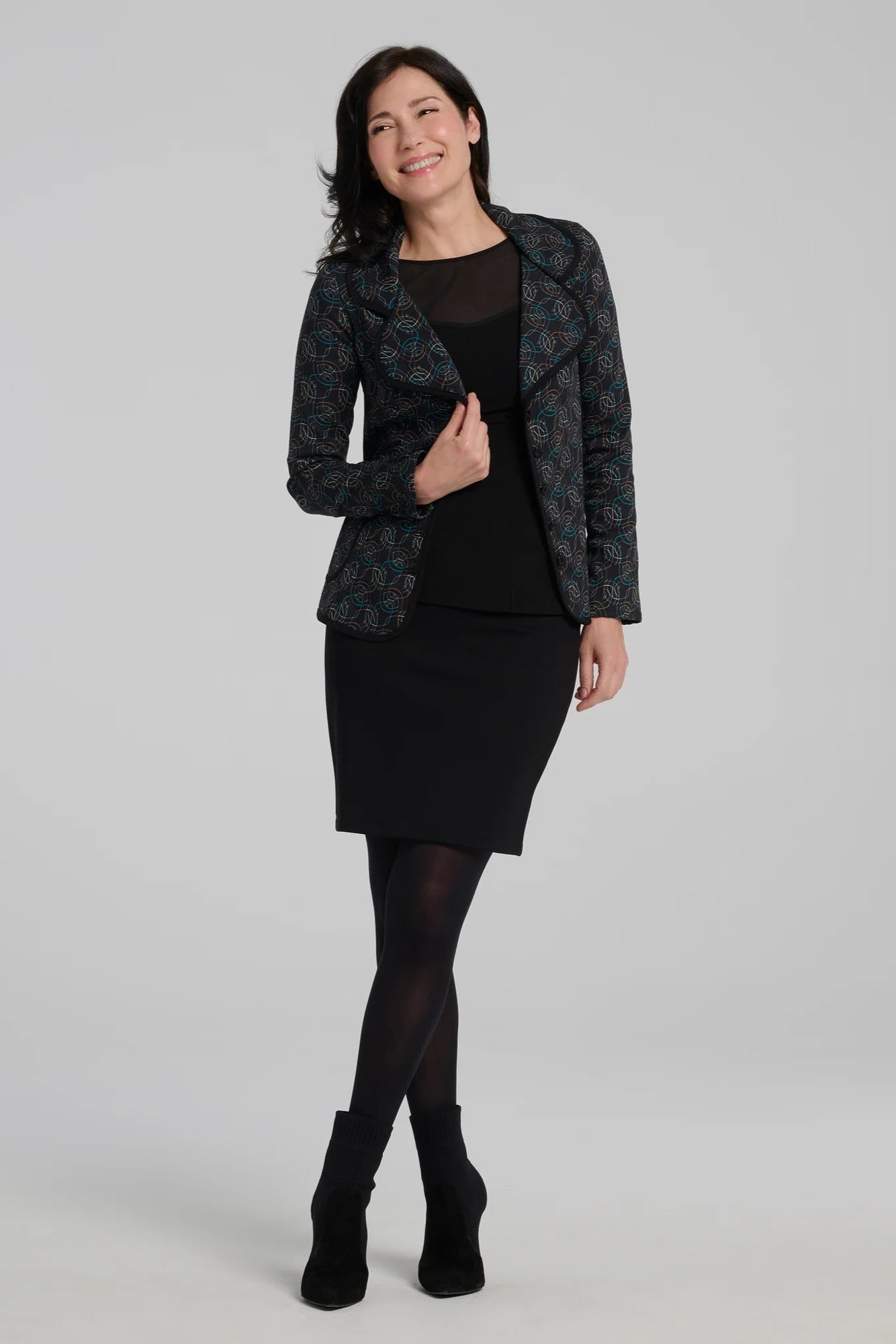 A woman wearing the Nara Jacket by Kollontai in Turquoise, a blazer with a turquoise on black geometric print, wit a rounded lapel collar, slim fit, and side pockets. She is wearing it with a black top and a black skirt and standing in front of a grey background. 