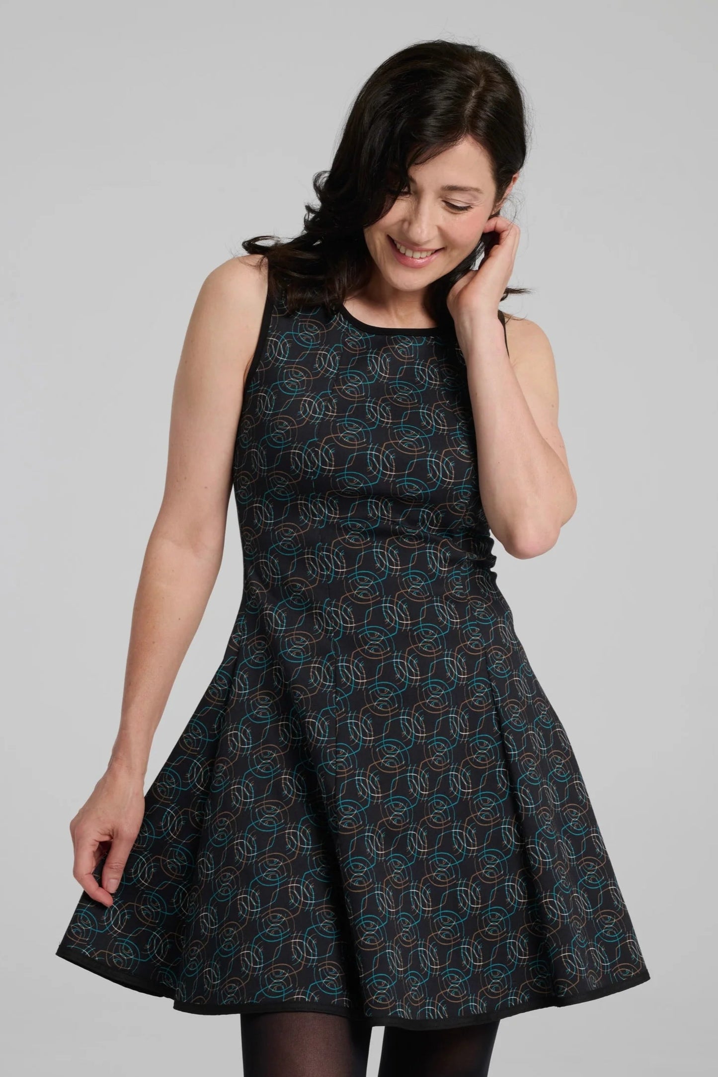 A woman wearing the Silba Dress by Kollontai in a Turquoise on black print, a sleeveless dress with a round neck, fit and flare shape, and above the knee hem. She is standing against a grey background. 