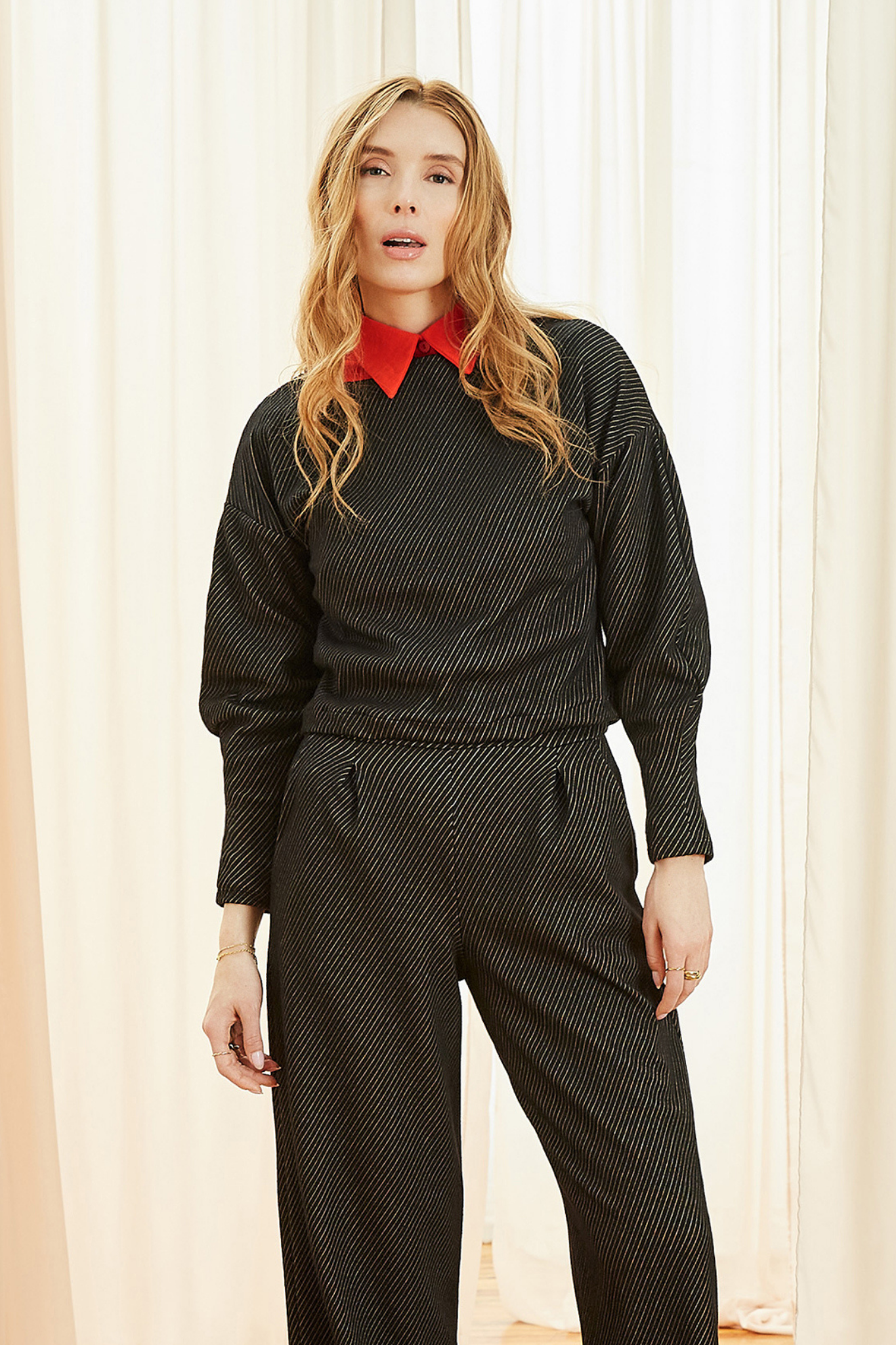 A woman wearing the Lewis Top by Melow in Black, featuring a diagonal stripe texture, boat neck, long puffed sleeves that snug at the wrists, and a cropped length. She is wearing it with matching pants and standing in front of a curtain. 