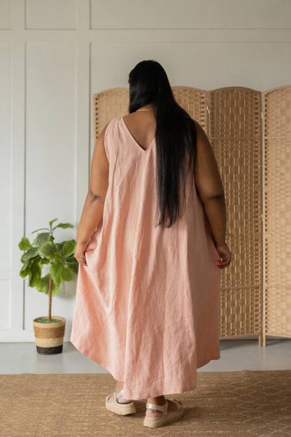 Back view of a woman wearing the Caroline Dress by Lights of All in Ballet Pink, a sleeveless dress linen maxi dress with a V-neck at the back. She is standing against a white wall with a woven screen behind her. 